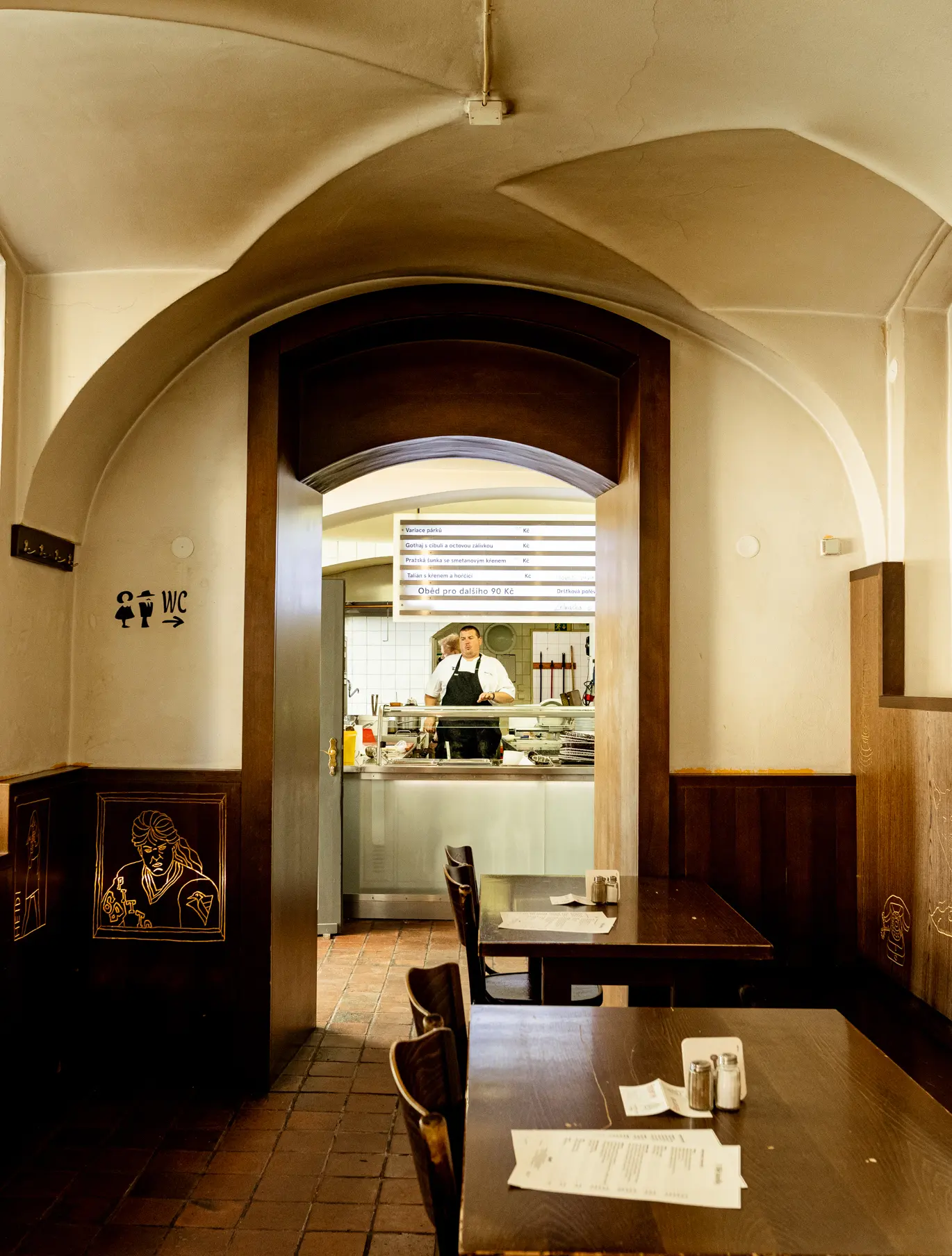 Interiér restaurace Lokál U Bílé kuželky s klenutým stropem, otevřeným průhledem do kuchyně a tradičními dřevěnými stoly.